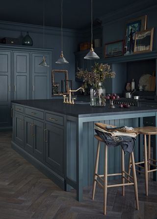 A dark gray-green kitchen with gold taps, pendant lighting and wooden bar stools.
