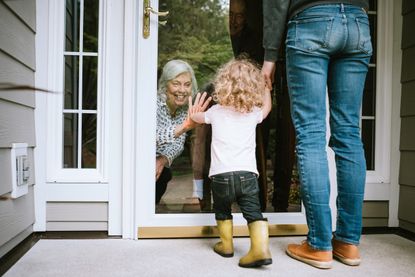 A little girl holding onto her mother's hand and saying hello to her grandmother inside her home. They are following safe social-distancing measures.