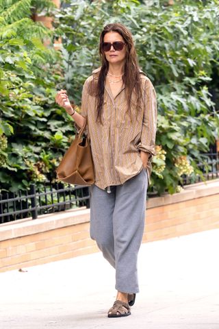 Katie Holmes carries a brown Khaite bag with a brown and white striped button-down shirt, gray sweatpants, fisherman sandals, and brown sunglasses.