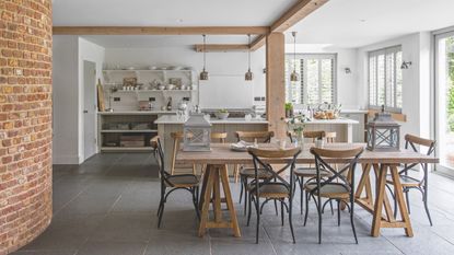 rustic kitchen with white walls and ceiling, open plan shelving, beams, large kitchen island, rustic table with wooden chairs, slate grey floor, shutters at window, metallic pendant lights, lanterns