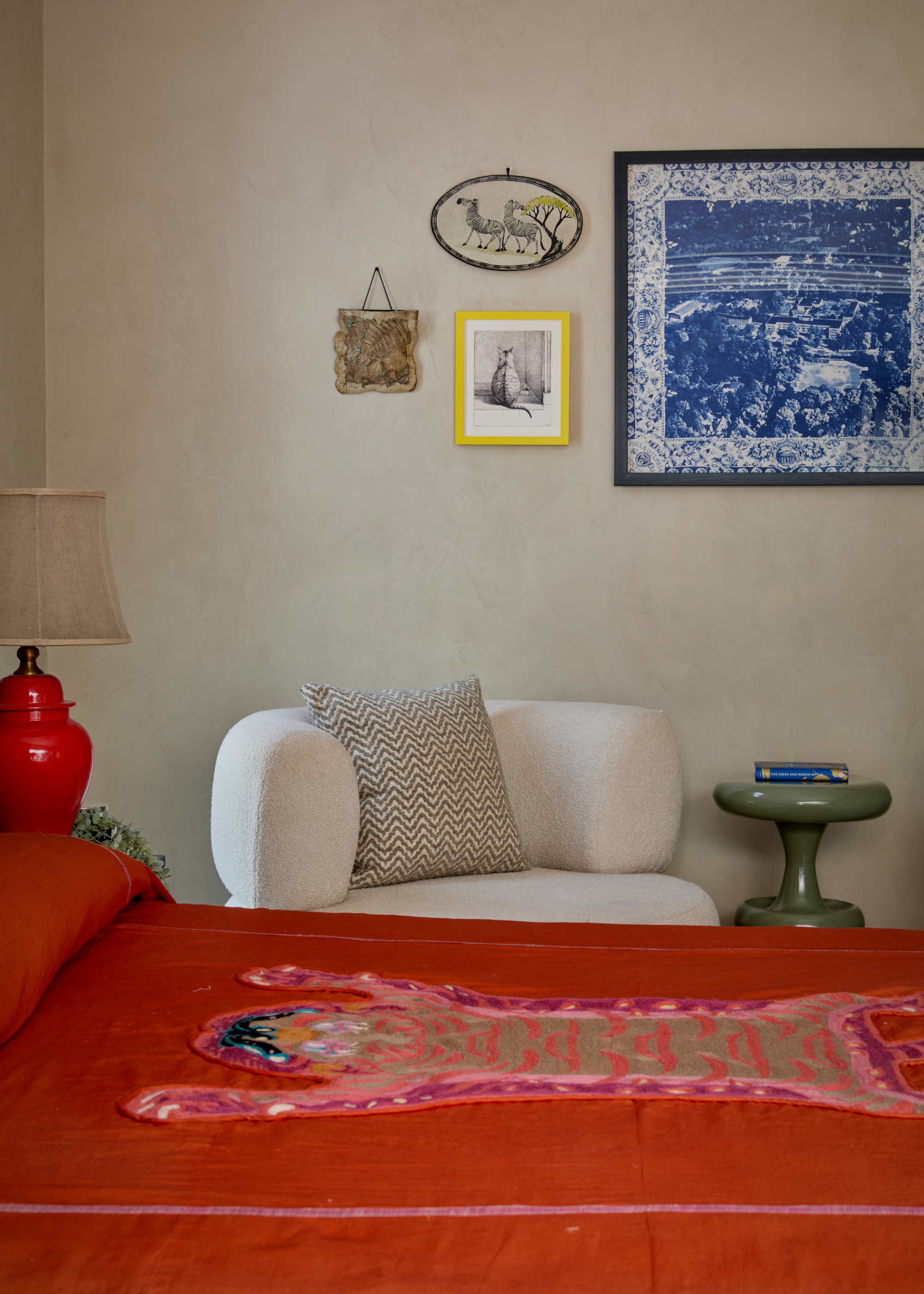 A summery bedroom with bright bedding, a sofa seater, a green side table, a bright lamp and a framed art gallery
