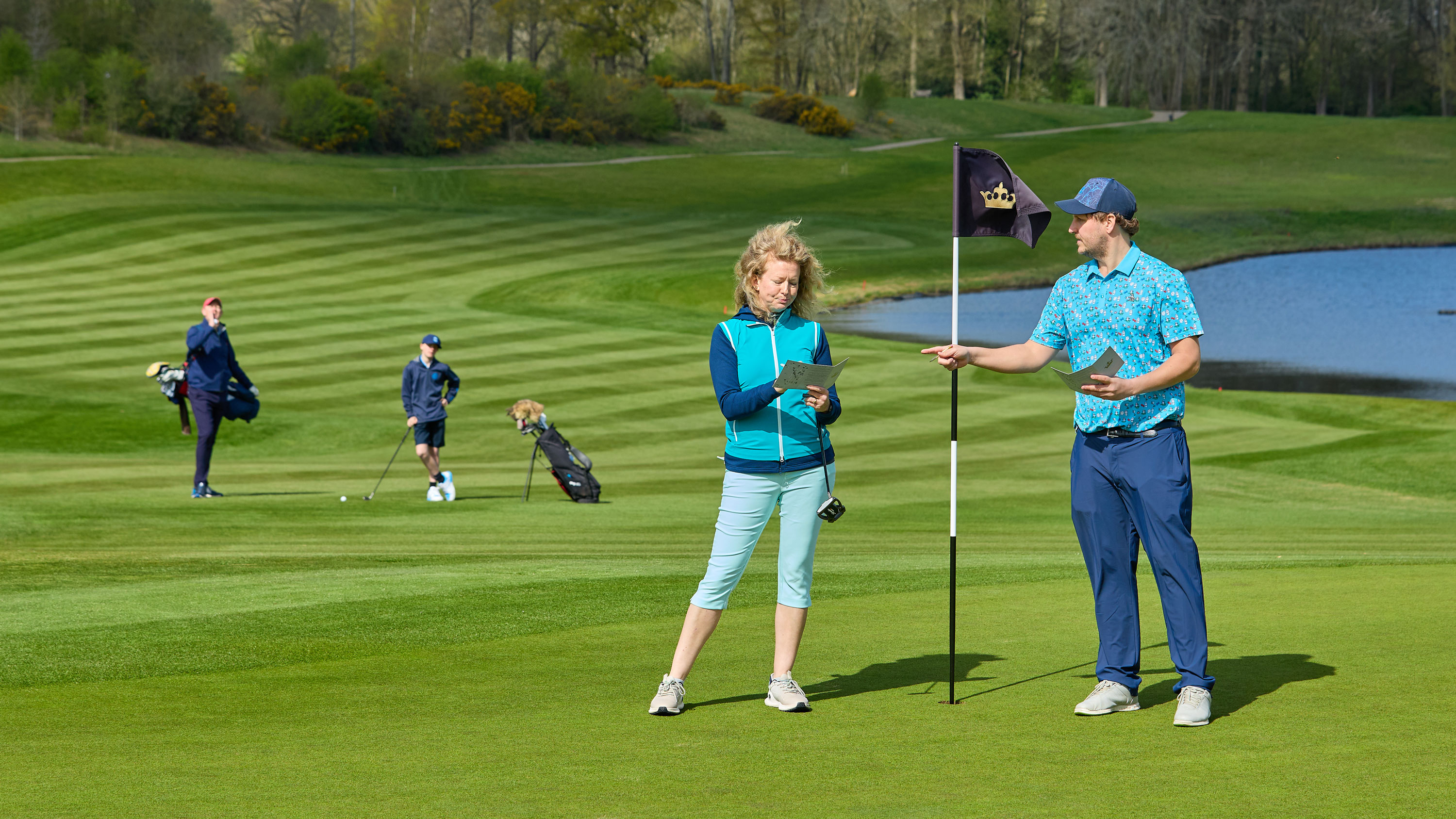 two golfers marking their scorecards on the green while two other golfers wait for them on the fairway behind