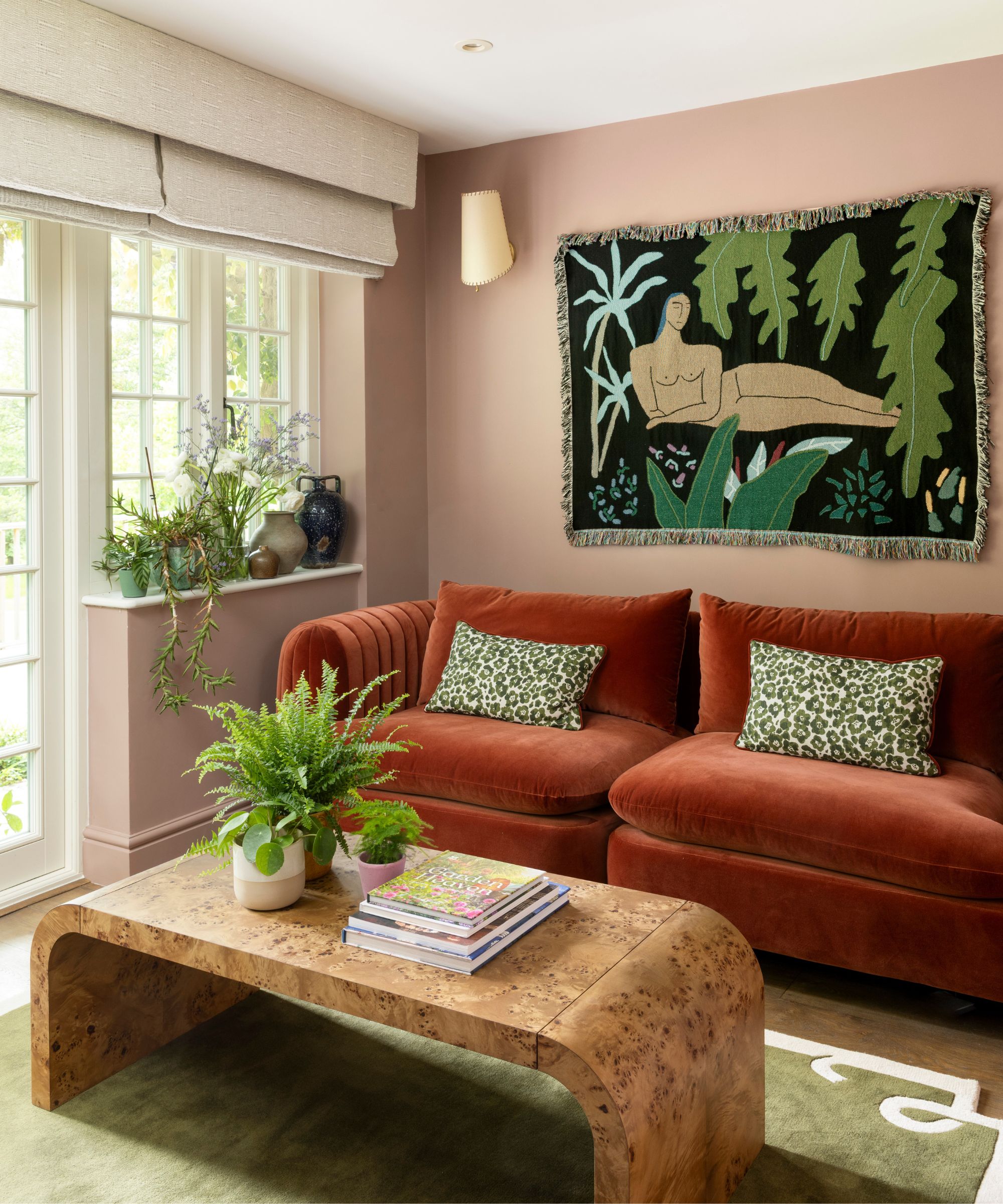 a garden room with dusty pink walls, a rust red velvet couch, blanket wall hanging, burl wood coffee table and sage green rug