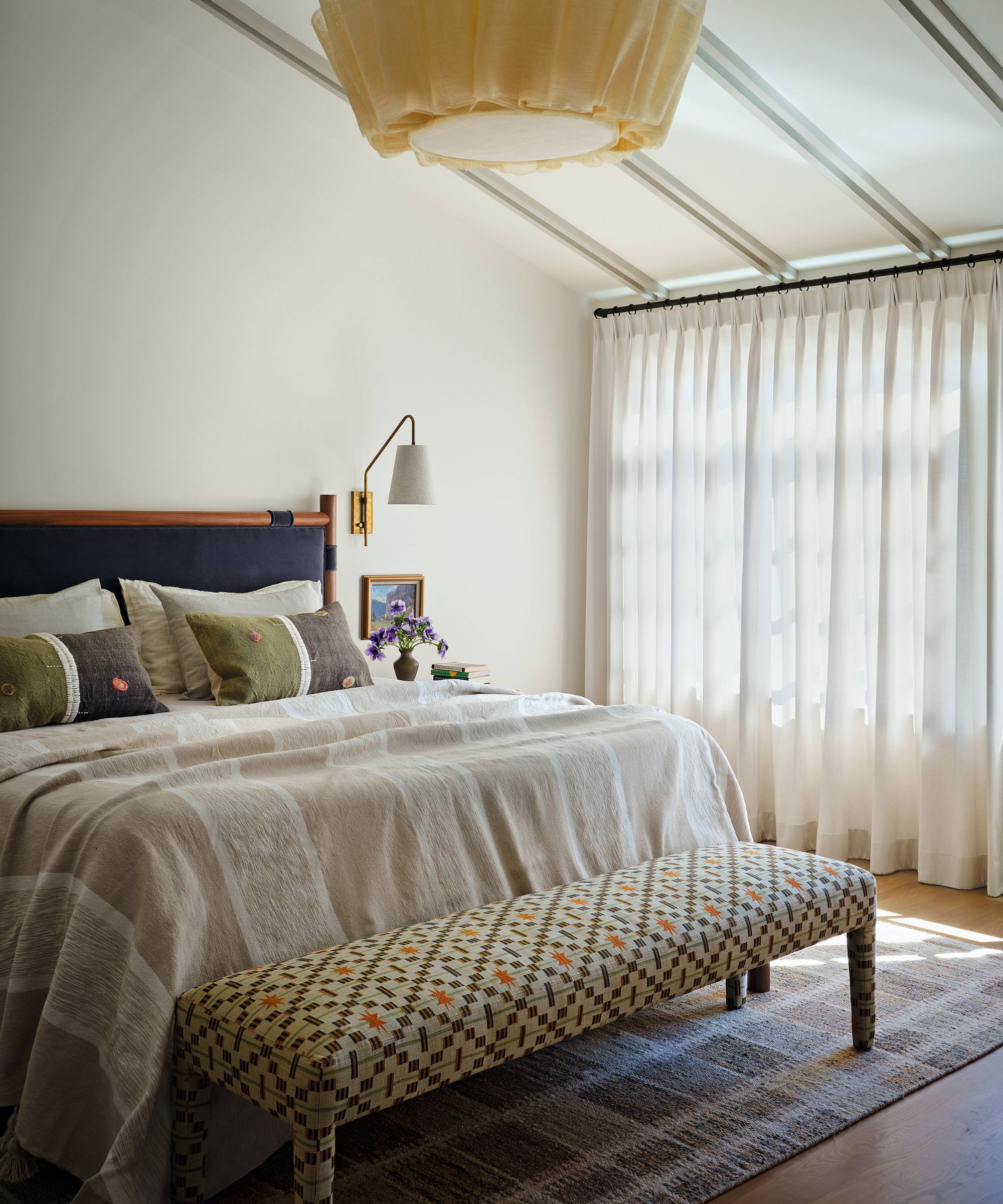 a neutral bedroom with vaulted ceiling with a wooden and navy bed and a patterned ottoman at the foot