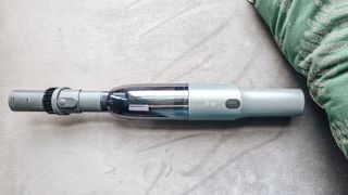 A gray handheld Brigii vacuum with a long handle and brush on top of a gray velvet chair, with a green and gold pillow next to it