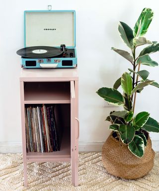 Pink metal Mustard locker, tall plant in woven basket, natural textured beige carpet, blue record player on locker, colorful wall art.