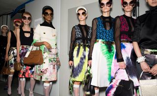 Models wear patterned skirts and jacket with black see through tops with sunglasses and hats