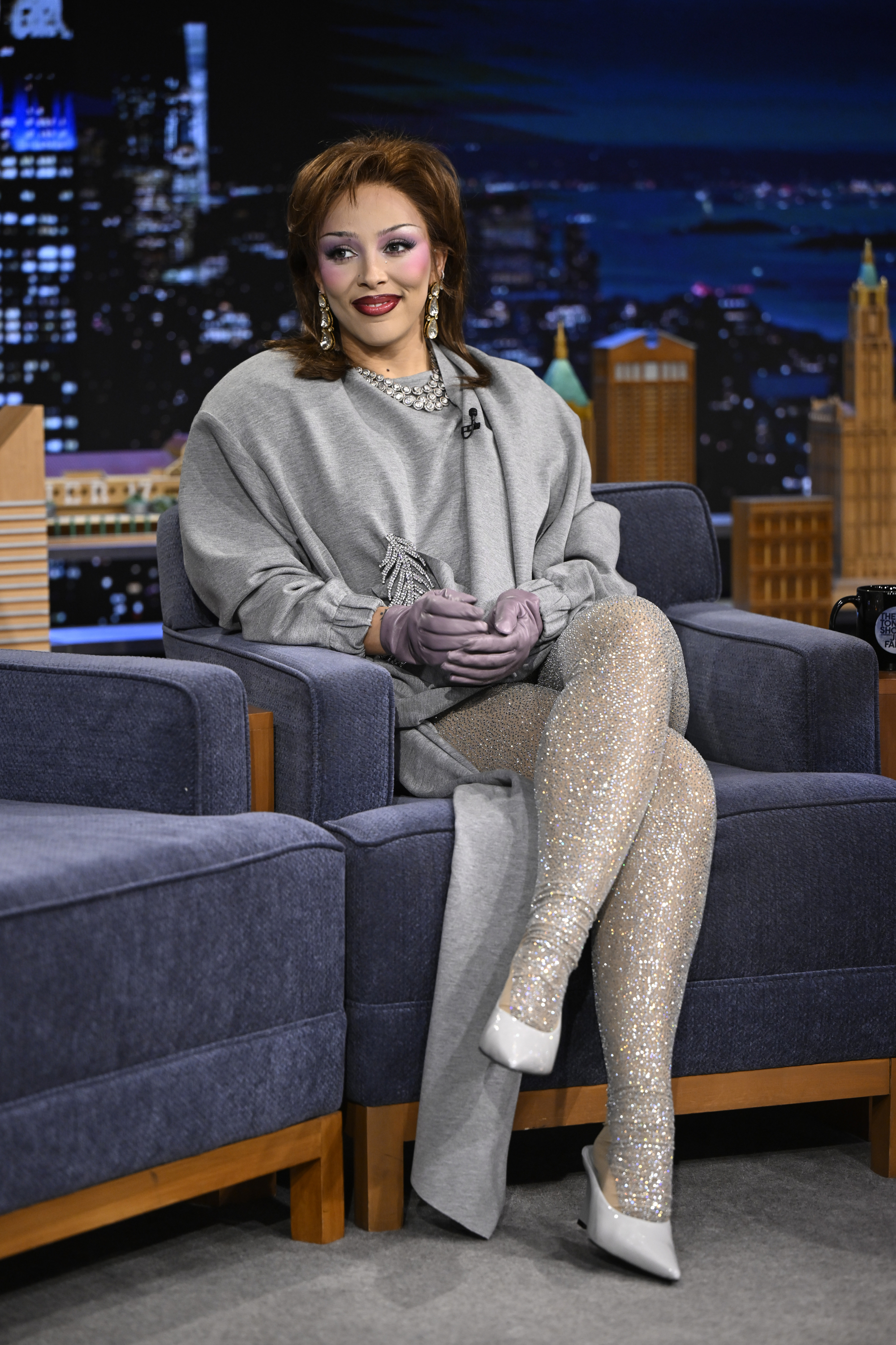 a woman with brown hair sitting on a gray couch on a talk show set wearing a gray dress with sparkly gray tights and gray pumps