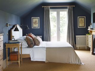 double bed with cream upholstered headboard and dark blue walls sloping ceiling and french doors period style radiators vintage accessories and fern botanical prints sisal flooring