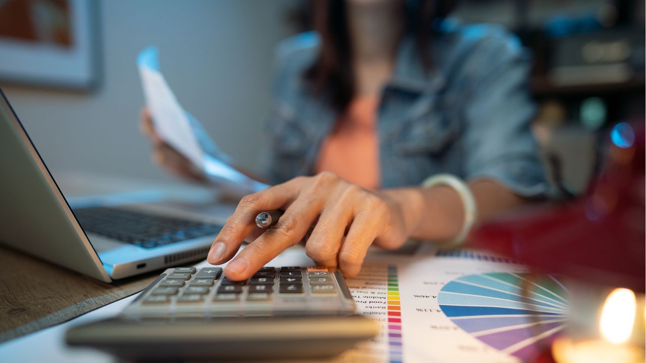 A woman uses a calculator next to her laptop, only her hand showing.