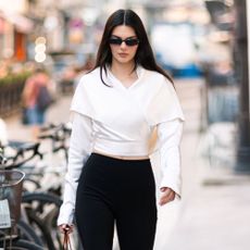 Kendall Jenner walks down the streets of Paris wearing a white wrap top, black high-waisted capri pants and black peep-toe heels. She hold a small black handbag in her clutches as accessories with black sunglasses.
