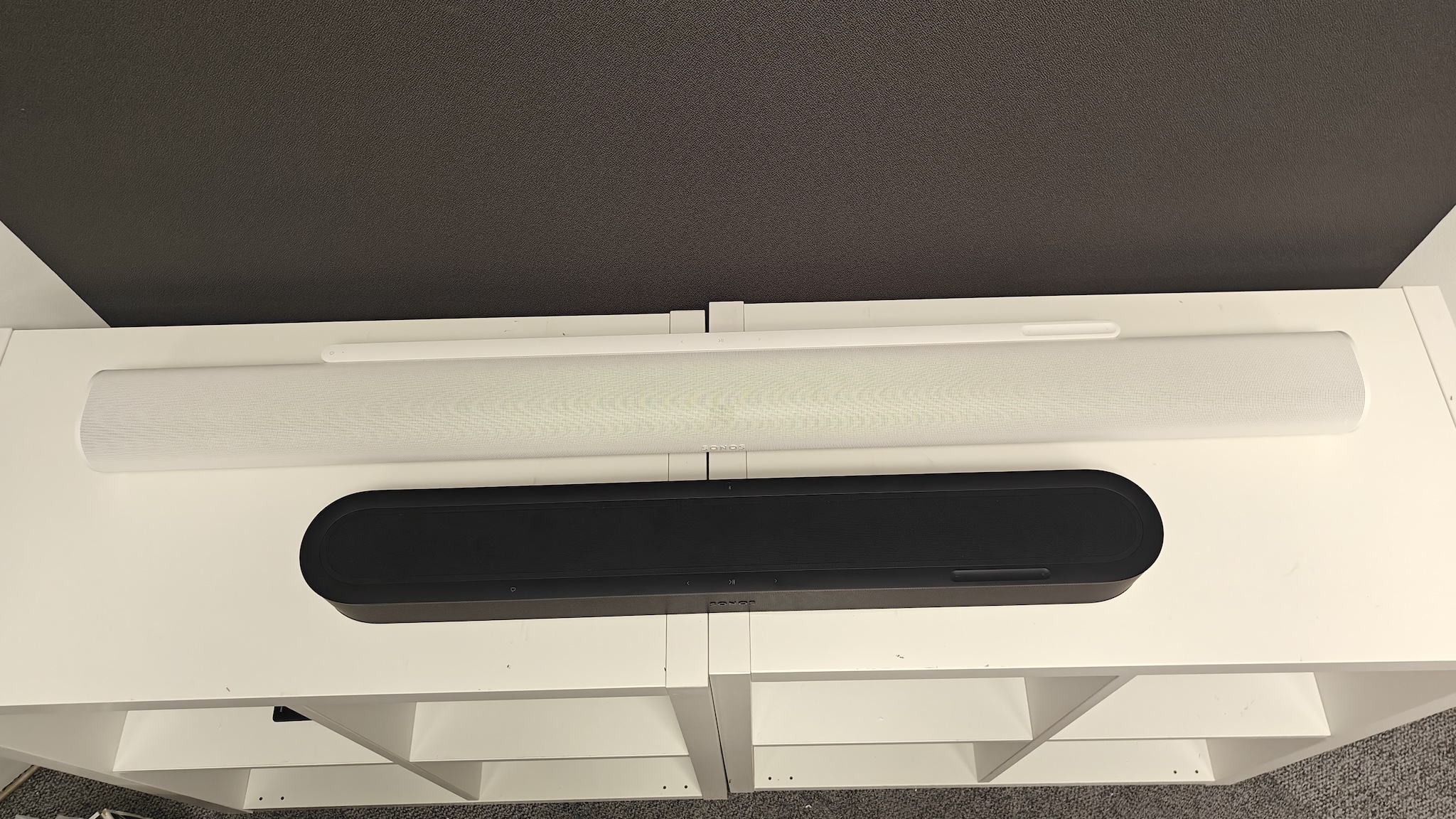 A black Sonos Beam Ultra and white Sonos Arc Ultra next to each other on a white TV cabinet, shot from above.