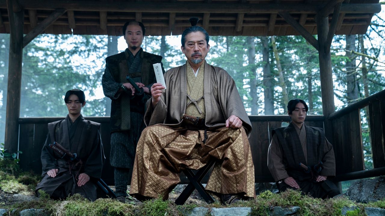 a man in gold japanese period wear (Hiroyuki Sanada as Yoshii Toranaga) sits while holding up a white letter, with one man standing behind him and two more kneeling at each side, in &#039;shogun&#039; (2024)