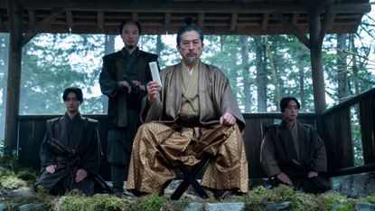 a man in gold japanese period wear (Hiroyuki Sanada as Yoshii Toranaga) sits while holding up a white letter, with one man standing behind him and two more kneeling at each side, in 'shogun' (2024)