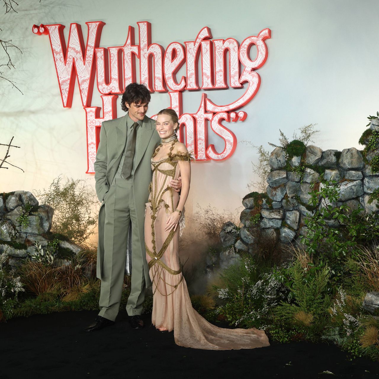 Jacob Elordi and Margot Robbie stand in front of a crumbling stone wall and greenery. The words "Wuthering Heights" are spelled out in red and white.