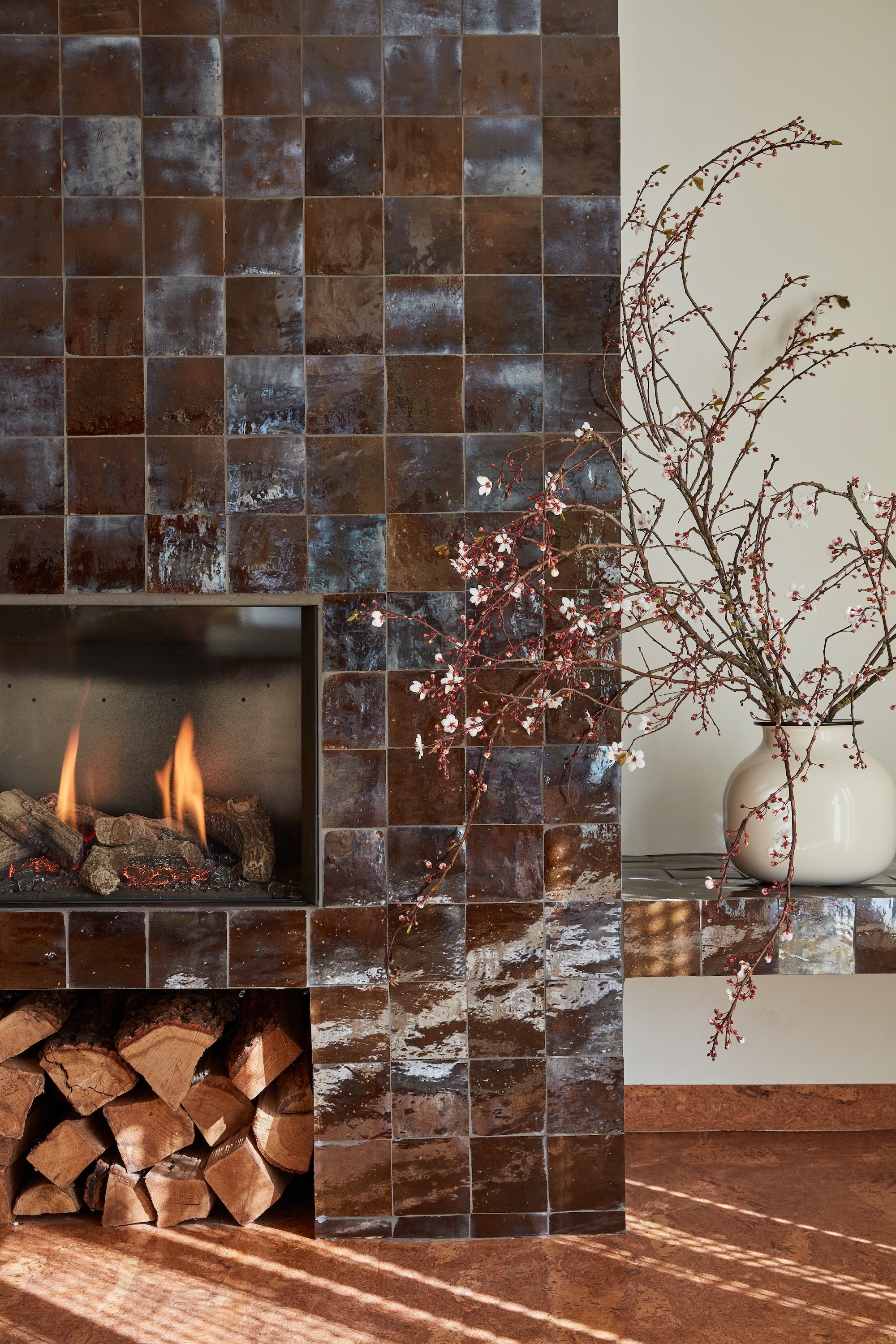 A seventies brown tiled fireplace with built in shelf to the right and a splay of natural blossom in a large vase