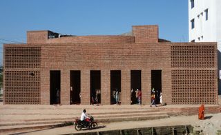 The facade of the Baitur Rauf Jame Mosque is built out of red brick. The Mosque is rectangle-shaped with multiple openings through which people are coming through.