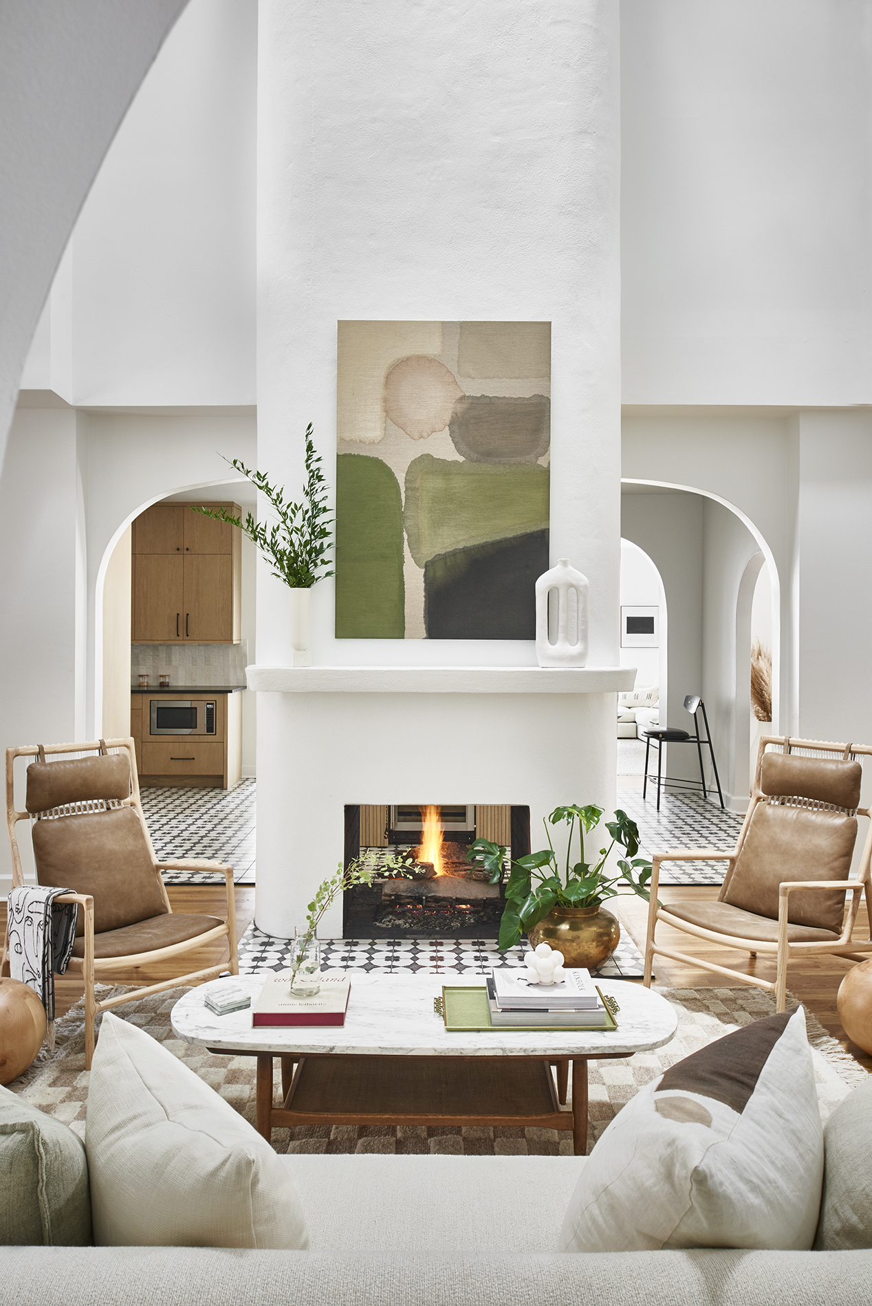 Image of a white minimalist living room with high ceilings. There is a fireplace in the middle with a green abstract painting hanging over it. There are two wooden and leather accent chairs around a white marble coffee table and a white sofa.