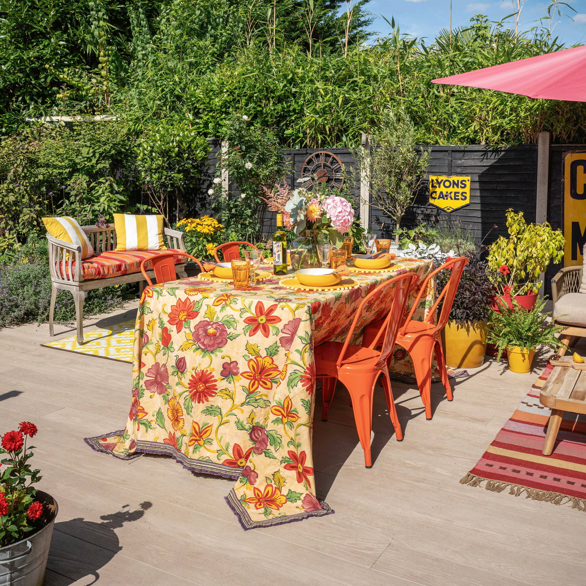 a garden with a black painted fence with a vintage sign with lots of planters filled with plants and flowers beside a dining table and chairs covered in a brightly coloured floral tablecloth