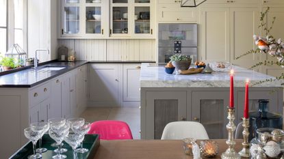 A bright and open-plan kitchen with white panelled walls, white cabinetry, black marble countertops, and a large island with marble countertop. In the foreground is a wooden dining table with a tray of coupes, lit red taper candles, and various decorations.