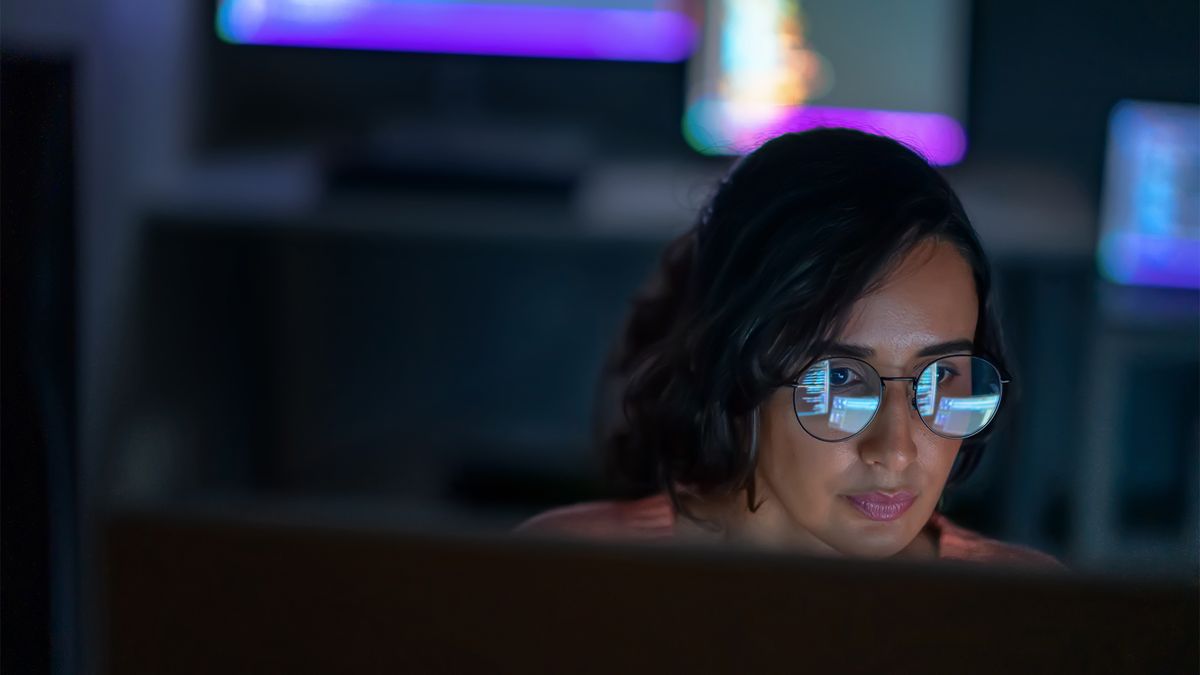 Female data analyst using AI tools on a desktop computer in a dark office space, with light from screen reflecting on spectacles. 