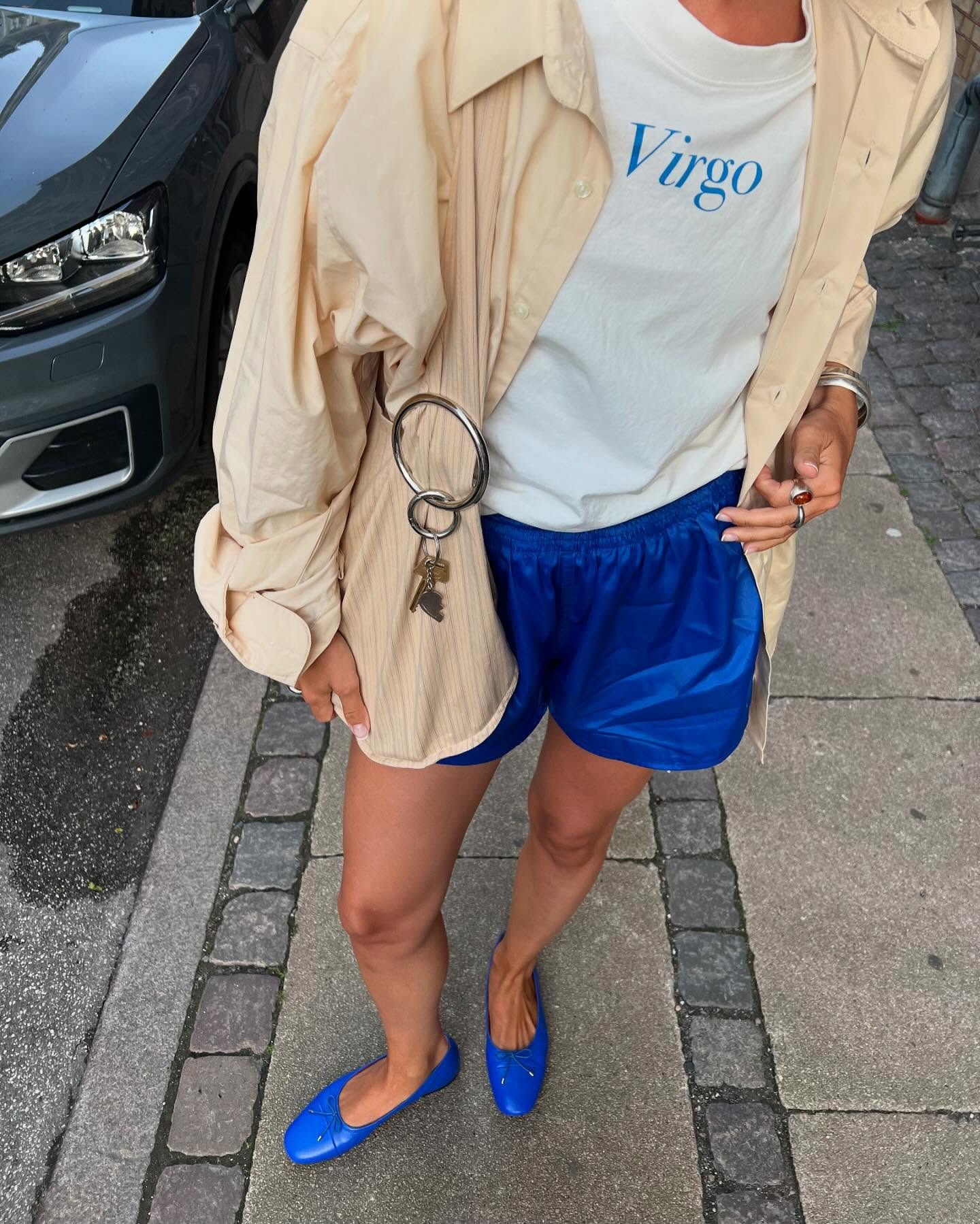 Danish style influencer Amalie Nielsen poses on a Copenhagen sidewalk wearing a chic 2026 outfit with a beige jacket, Virgo graphic T-shirt, bright blue shorts, and royal blue ballet flats