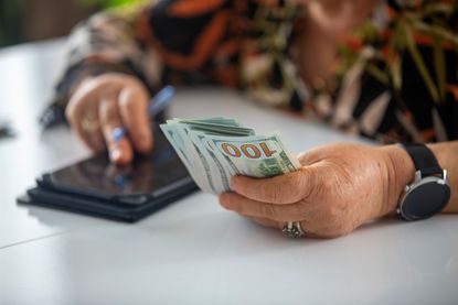 A senior woman holds a handful of cash while doing math on her phone's calculator.