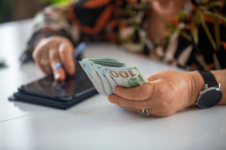 A senior woman holds a handful of cash while doing math on her phone's calculator.