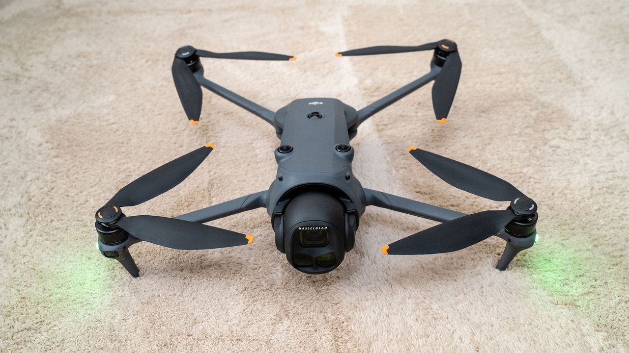A grey drone has four propellers extending out of each corner of the central body with green lights shining from the front two propellers on a beige carpet.