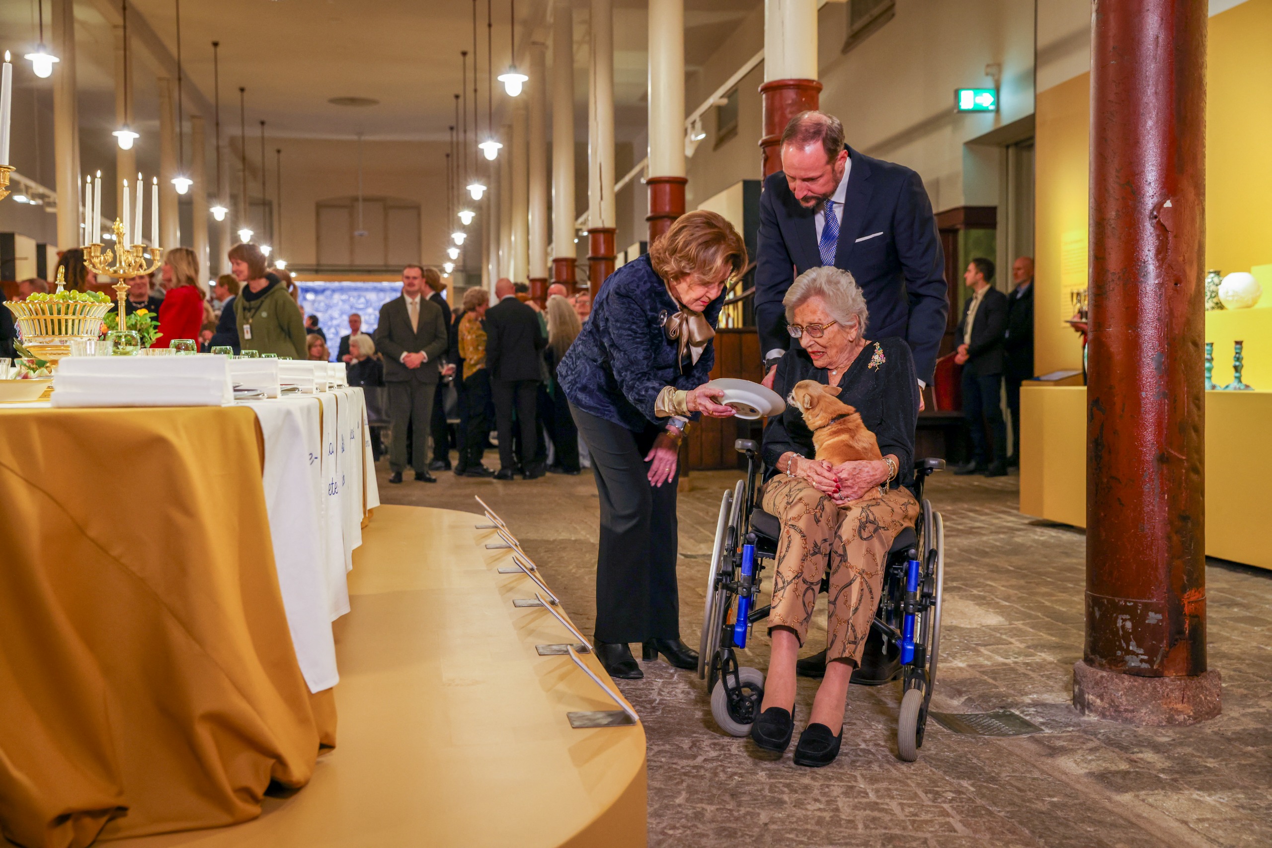 Queen Sonja leaning over to show a plate to Princess Astrid and her dog