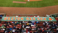 The Boston Red Sox logo on top of the dugout box at Fenway Park, viewed from above and shot with a telephoto lens. The crowd are immediately before it, with the green baseball diamond visible at the top of the frame.