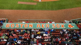 The Boston Red Sox logo on top of the dugout box at Fenway Park, viewed from above and shot with a telephoto lens. The crowd are immediately before it, with the green baseball diamond visible at the top of the frame.