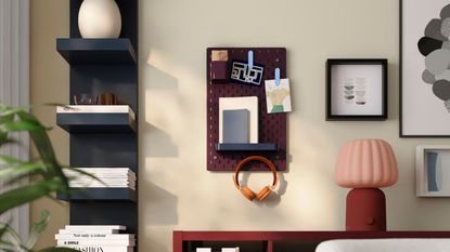 A burgundy pegboard with a shelf and pictures clipped onto it next to artworks and a vertical blue wall shelf and above a red unit with a red table lamp
