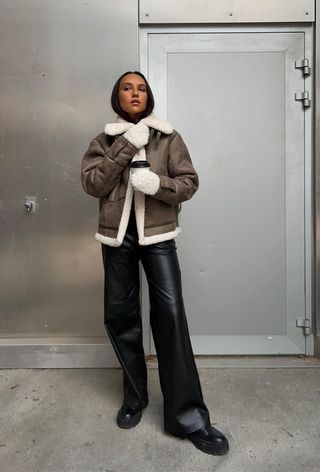 how to wear Chelsea boots is shown in a photo of a woman standing outside wearing a brown shearling jacket with black leather trousers and a black chelsea ankle boot