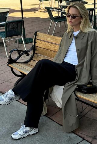 a photo showing what shoes to wear with wide-leg pants with a woman sitting on a bench wearing a long tan trench coat over a white t-shirt with black wide leg trousers and white chunky sneakers