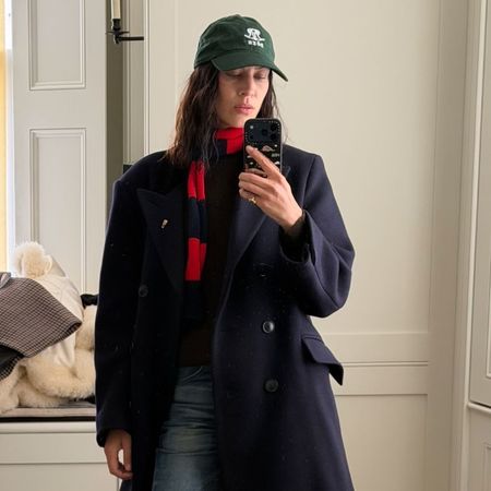 Alexa Chung takes a mirror selfie wearing a navy coat with stove pipe jeans, Tabi loafers and a green cap.