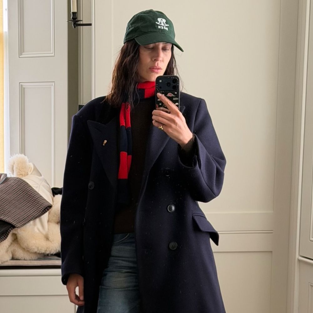 Alexa Chung takes a mirror selfie wearing a navy coat with stove pipe jeans, Tabi loafers and a green cap.