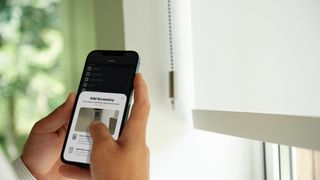 Someone holding a phone displaying the Eve for HomeKit app beside Eve Motion Smart Blinds.