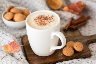 A pumpkin spiced latte in a white mug, with some cookies and cinnamon sticks.