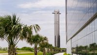 A massively tall chrome booster stands behind a row of palmettos next to a black, reflective building coverd in large windows.