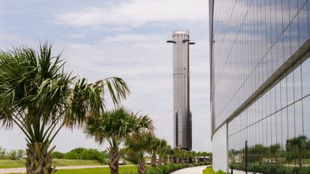 A massively tall chrome booster stands behind a row of palmettos next to a black, reflective building coverd in large windows. 