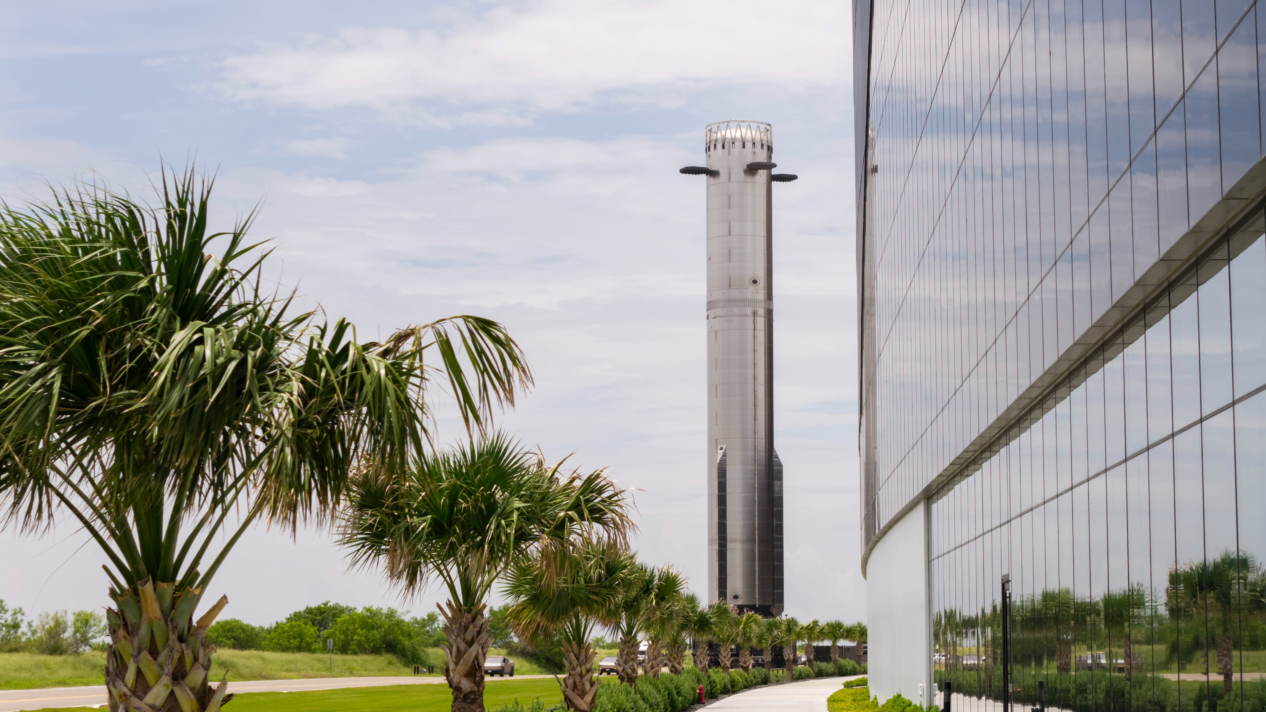A massively tall chrome booster stands behind a row of palmettos next to a black, reflective building coverd in large windows. 