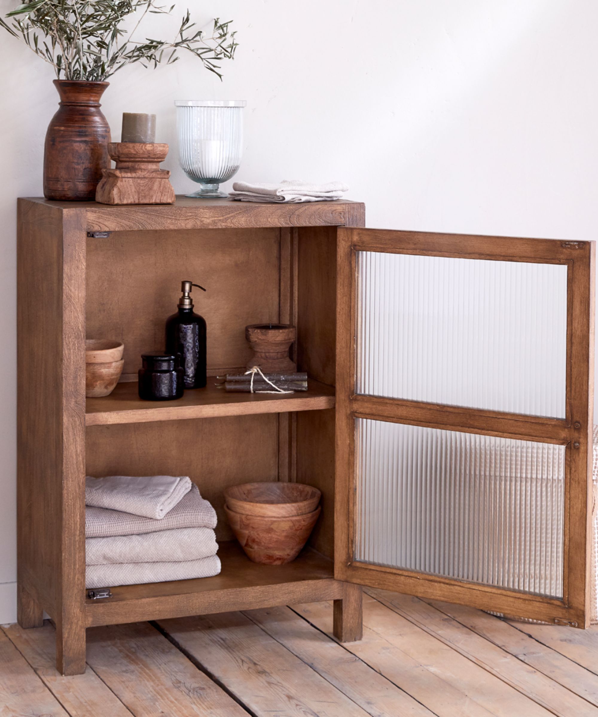 A compact wooden cabinet with reeded glass door stores folded towels and toiletries. Natural wood tones and simple ceramics create a warm, understated bathroom feel.