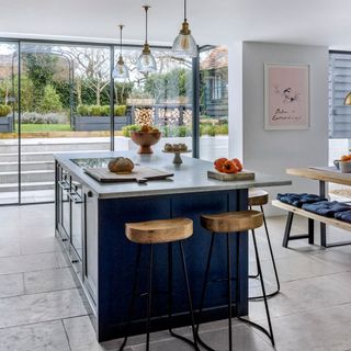 blue kitchen island with wooden stools, grey floor tiles and large back windows