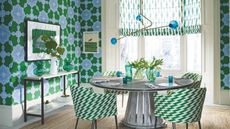 A green, blue and white dining area with green and blue patterned wallpaper, a round table with green and white chairs, a blue and gold statement light fixture, a white and brown console table with a white jug and green vases under a framed artwork, and large bay windows with green and white blinds.