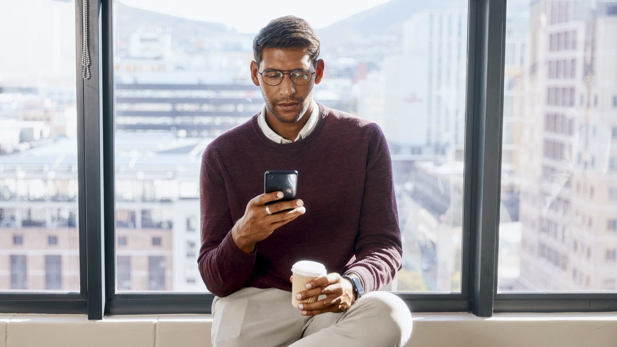 A young man sits at a window in an office building looking at his phone in surprise while holding a coffee. 