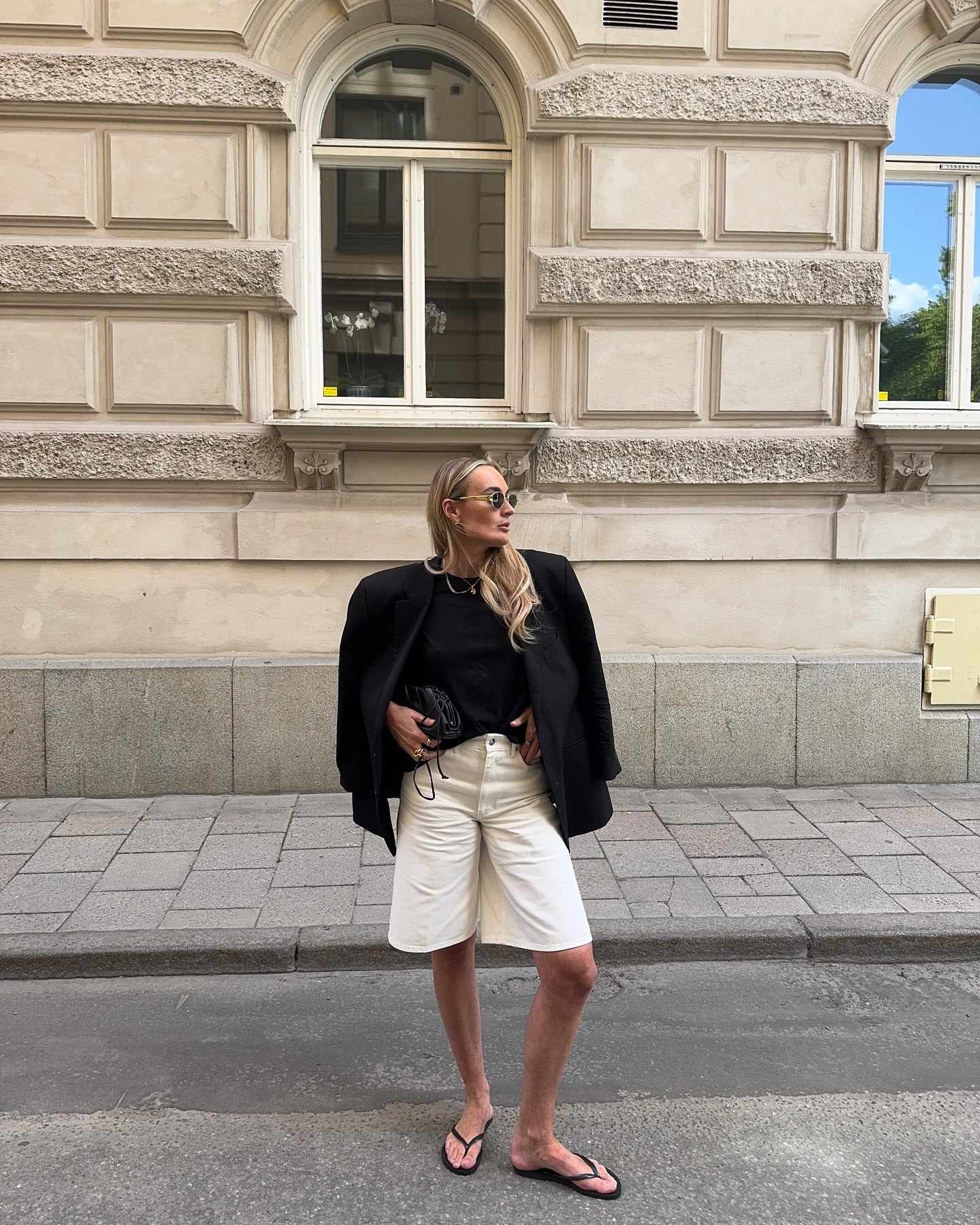 Woman wears black blazer, black T-shirt, white long denim shorts, black flip-flops