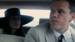 Julia Roberts sitting in the backseat of a car wearing a big black hat. Matt Damon is sitting in the car in front of her.