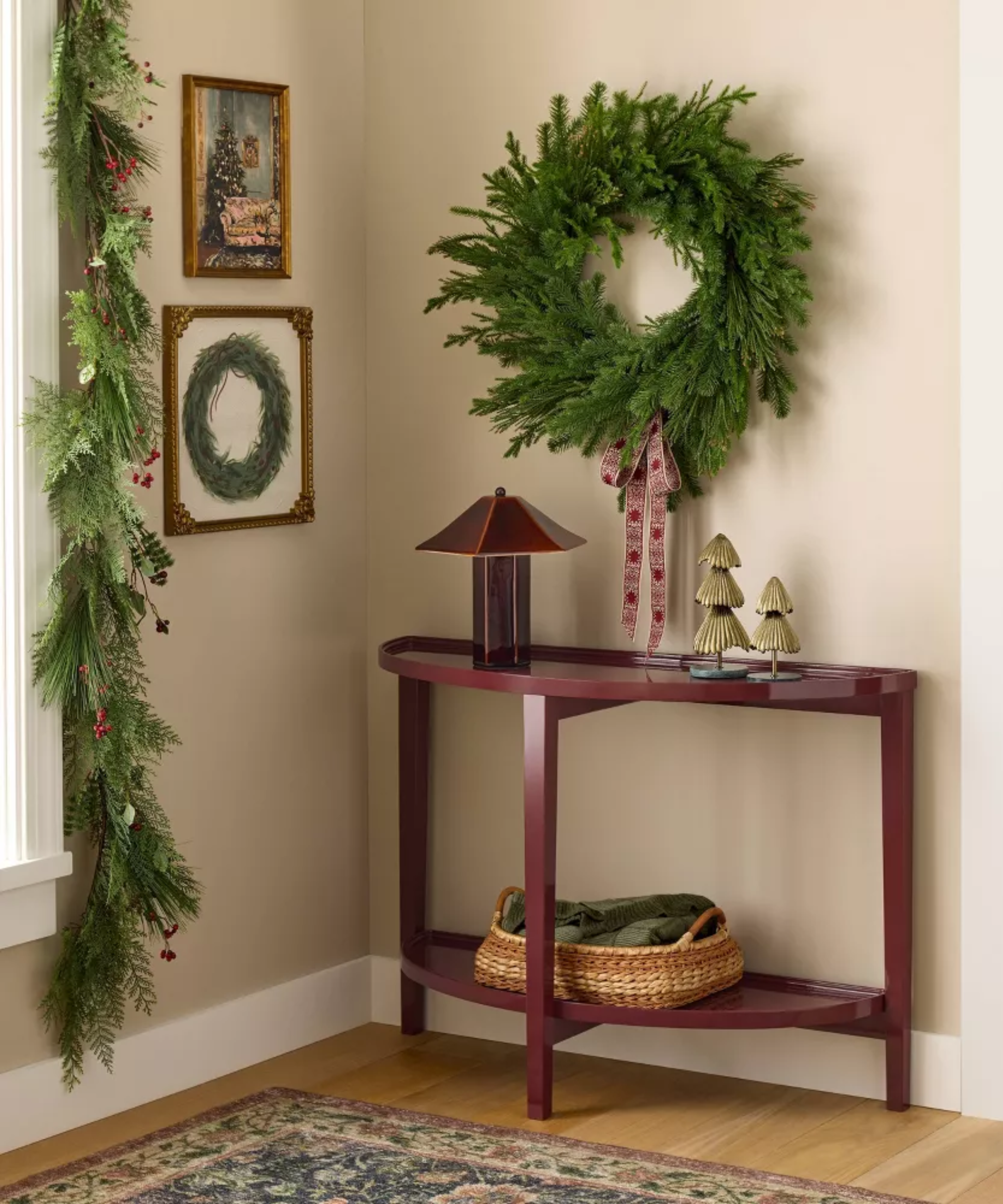 a corner of a room with a table decorated with some Christmas decor, a wreath hanging on the wall, two pictures hanging, and garland hanging around the window on the left side