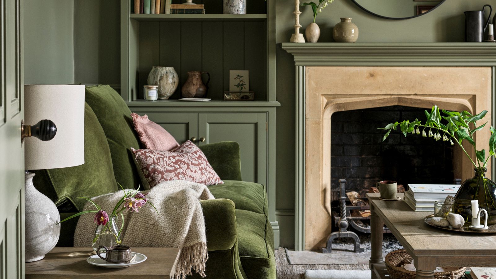 A green color-drenched living room with a deep green velvet sofa against the wall. Pink throw pillows. A light wood coffee table in front of it. 